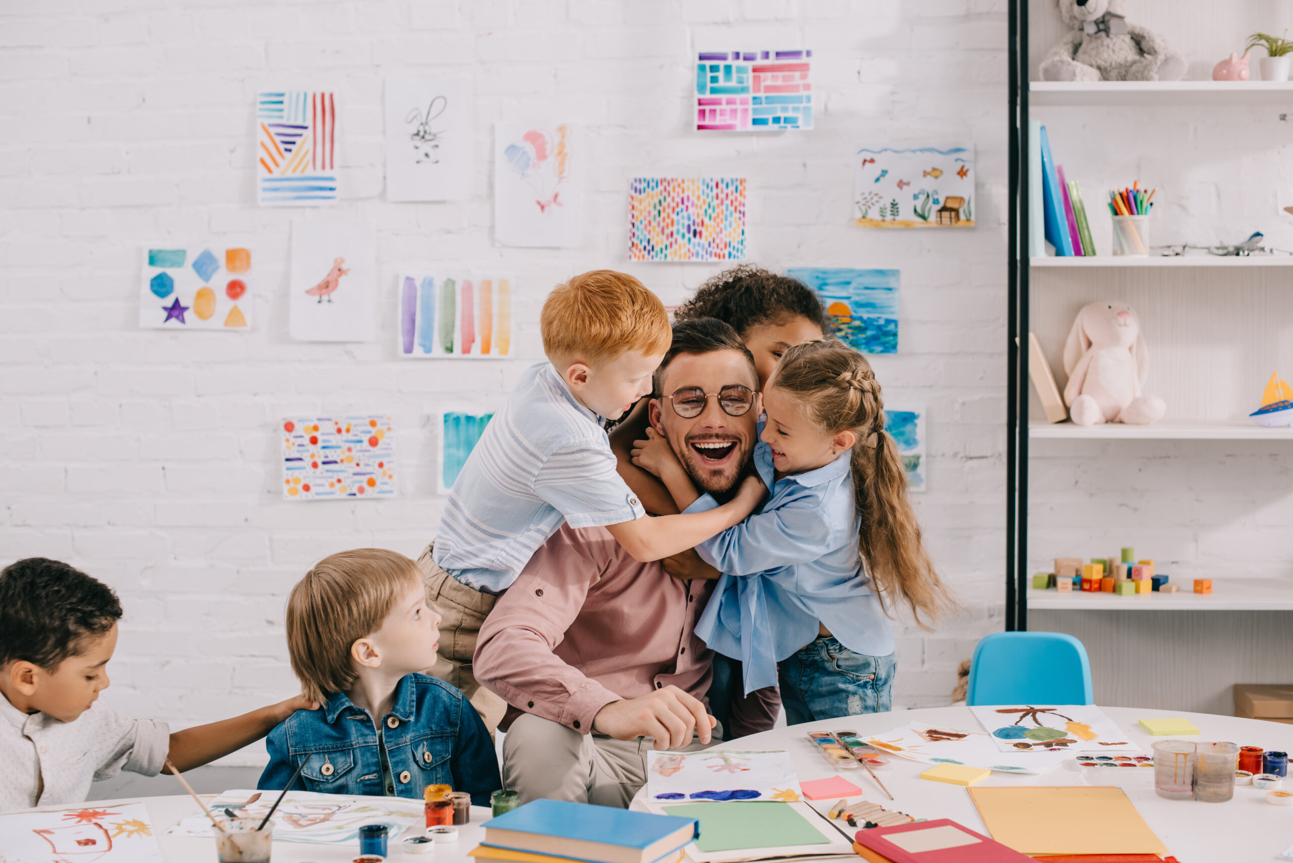 interracial kids hugging happy teacher at table in classroom